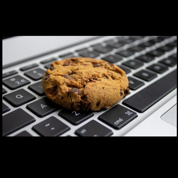 Un cookie aux pépites de chocolat repose sur un clavier d'ordinateur portable argenté. Le biscuit doré et croustillant contraste avec les touches noires et grises du clavier, évoquant un jeu de mots visuel entre les cookies informatiques et les cookies comestibles.