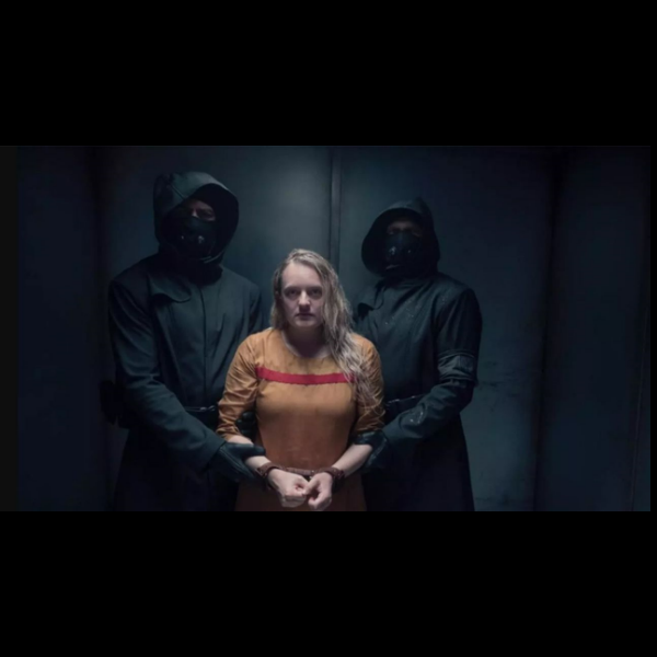 Dans un couloir étroit, sombre et métallique, une femme blonde en uniforme orange usé, marqué d'une bande rouge au niveau de la poitrine, se tient menottée, les poignets liés devant elle. Ses cheveux sont trempés, son visage fatigué, marqué par une tension visible, et son regard est dirigé droit vers l’avant, déterminé, presque accusateur. Elle est encadrée de chaque côté par deux hommes en tenue noire intégrale, masqués et gantés, portant des capuches qui obscurcissent leurs visages. Chacun tient fermement un de ses bras, soulignant la privation totale de liberté et le rapport de force brutal. L’éclairage est froid, limité, concentré sur la femme, accentuant le contraste entre la clarté qui révèle sa vulnérabilité et l’ombre dans laquelle se tiennent ses gardiens. L’ambiance évoque une scène d’interrogatoire, de détention ou de transfert vers une destination inconnue, dans un climat oppressant et autoritaire. Le décor dépouillé, sans repère, renforce le sentiment d’enfermement et de danger latent. L’image incarne visuellement l’abus de pouvoir, la surveillance constante, et la violence systémique exercée contre les femmes dans l’univers de La Servante écarlate.